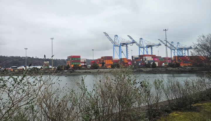 Four blue cranes with stacks of colorful shipping containers next to a waterway.