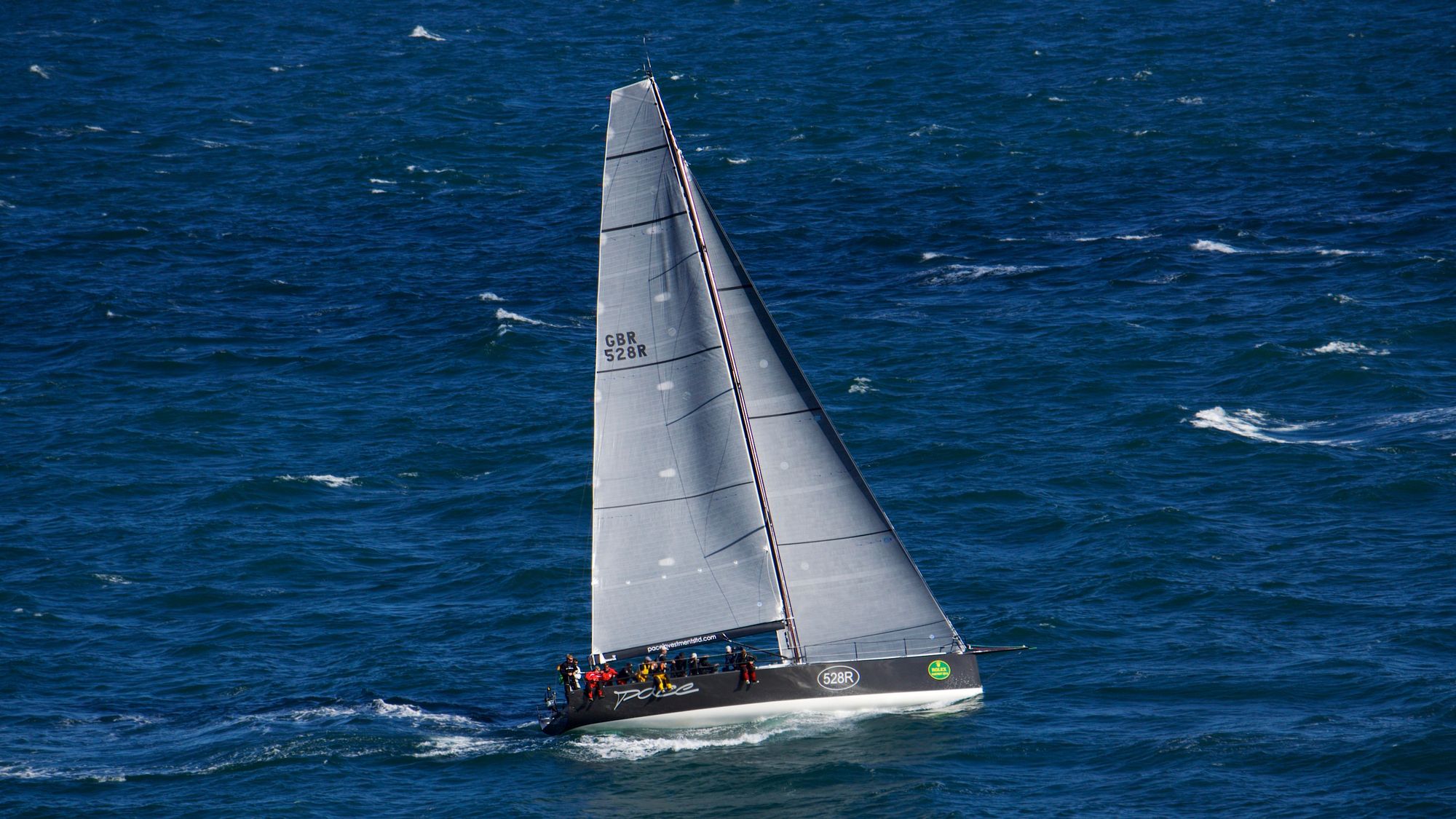 A sleek race boat with logos and the crew sitting on one side, sailing through blue water with whitecaps.