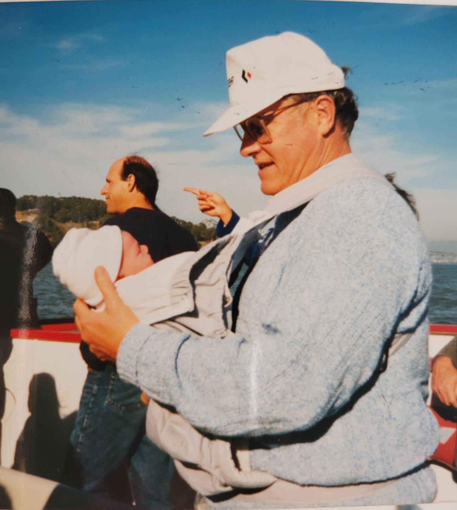 A man wearing glasses and a hat on the deck of a boat holds a small baby in a carrier on his chest.