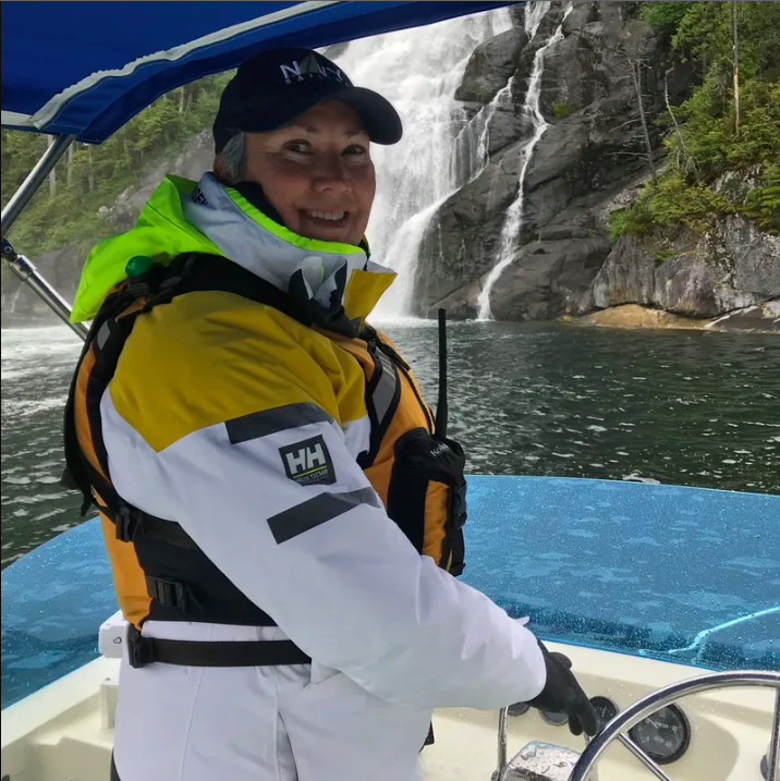 A woman in a lifejacket at the helm of a boat in front of a waterwall.