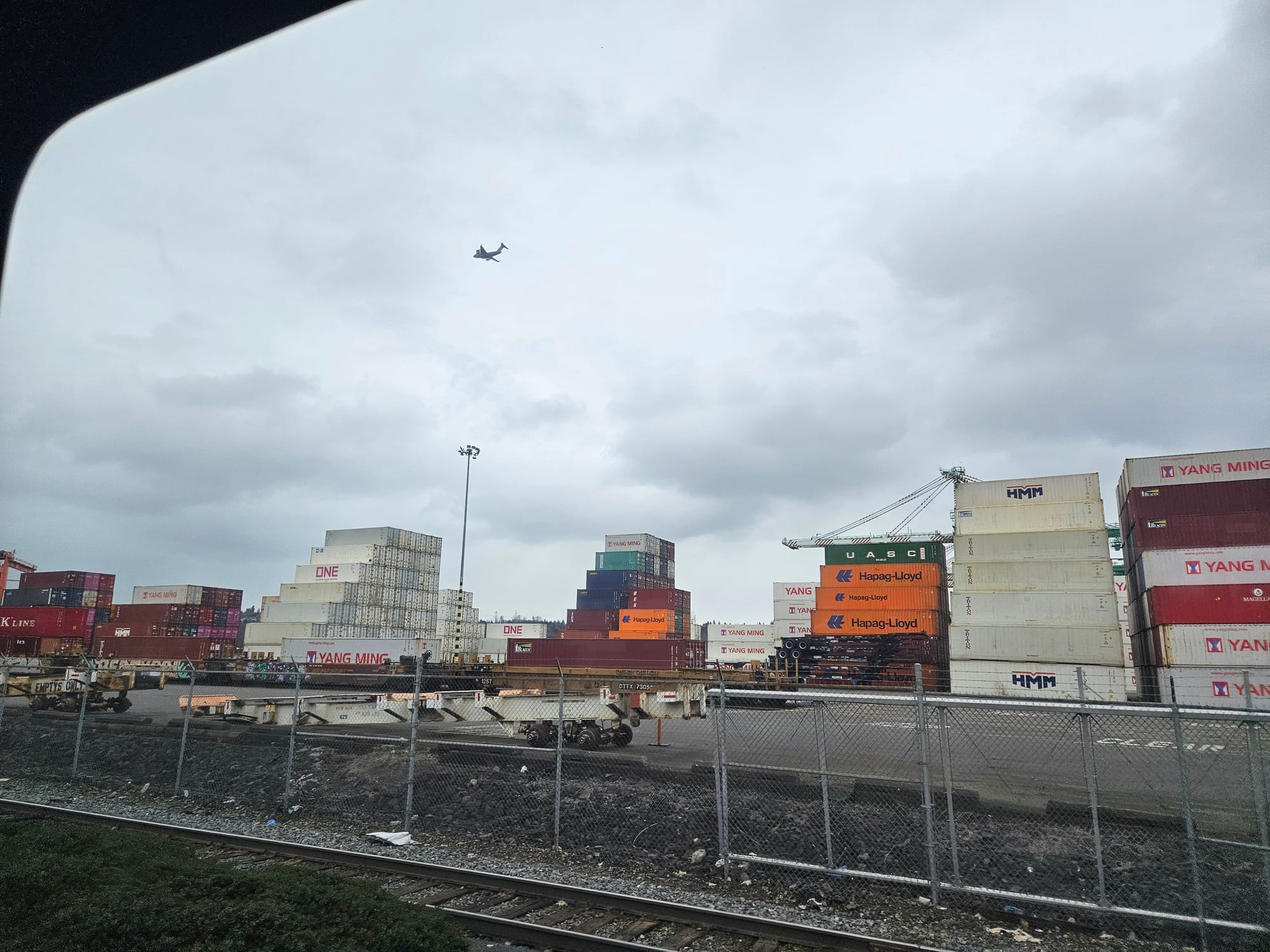 Large rectangular boxes stacked in a parking lot with train tracks and a fence in the foreground.