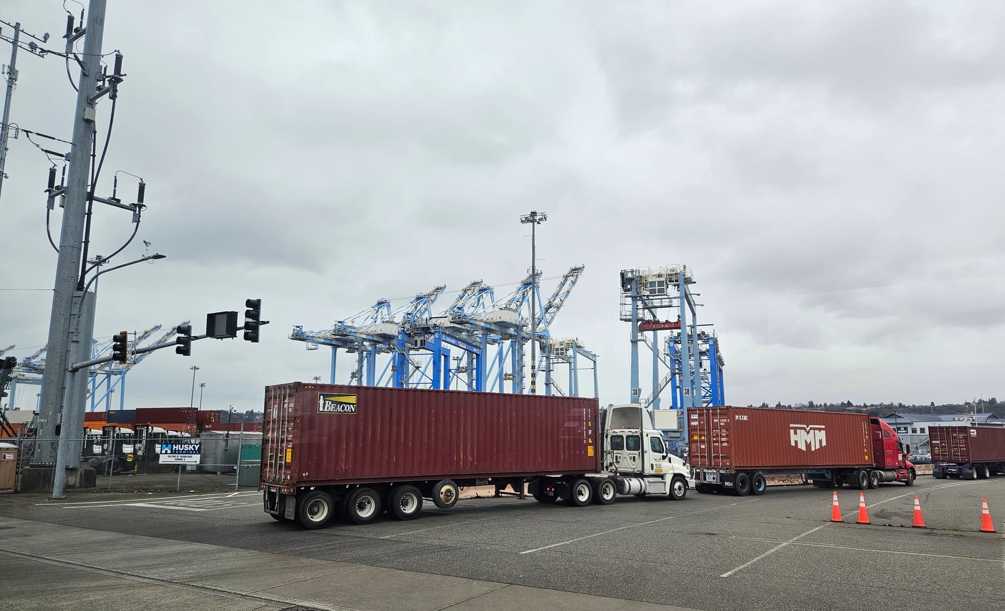Three trucks loaded with shipping containers wait in a line near an intersection, large blue cranes are in the background.
