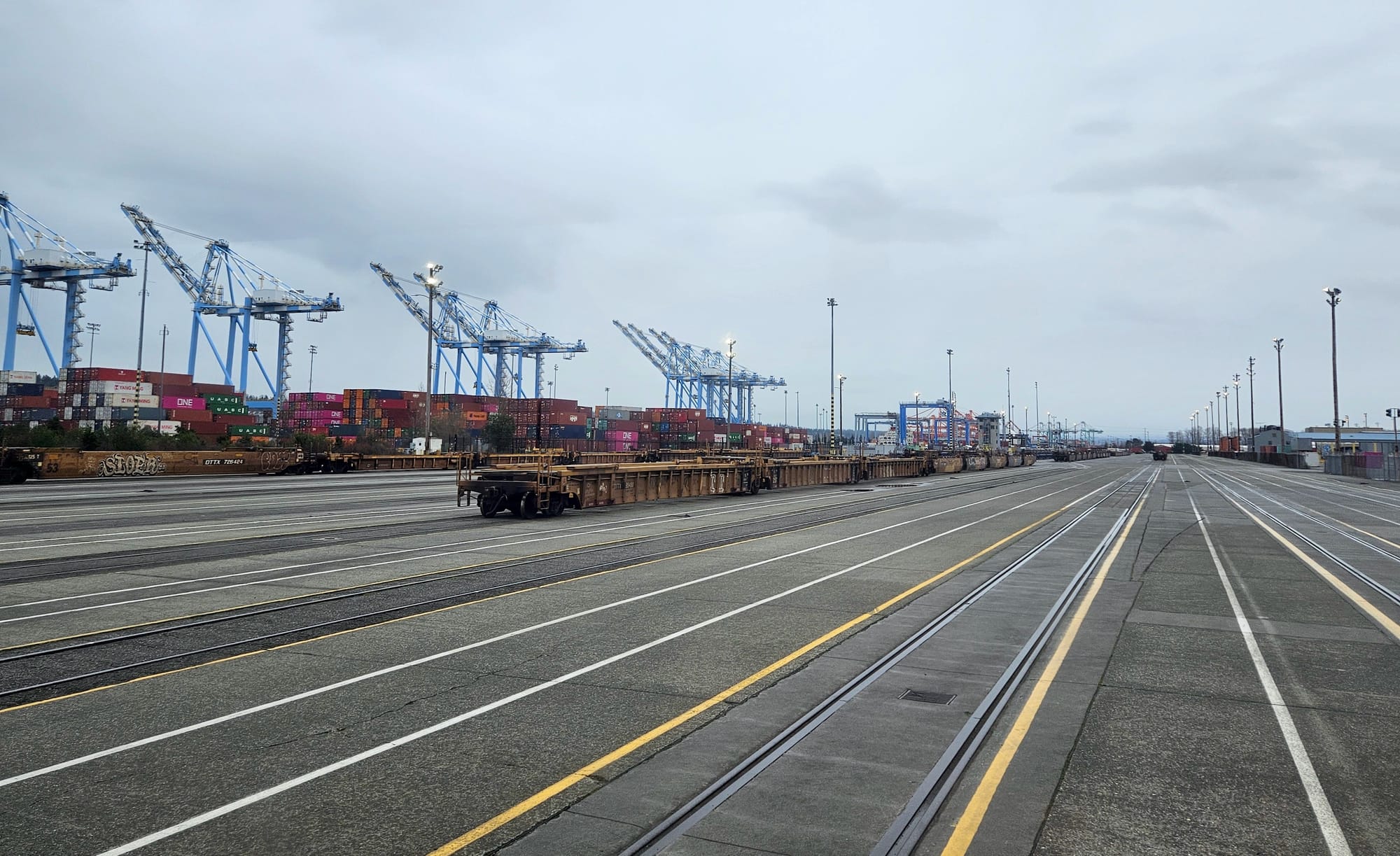 Half a dozen railroad tracks next to each other on a large concrete area with shipping containers and cranes in the background.
