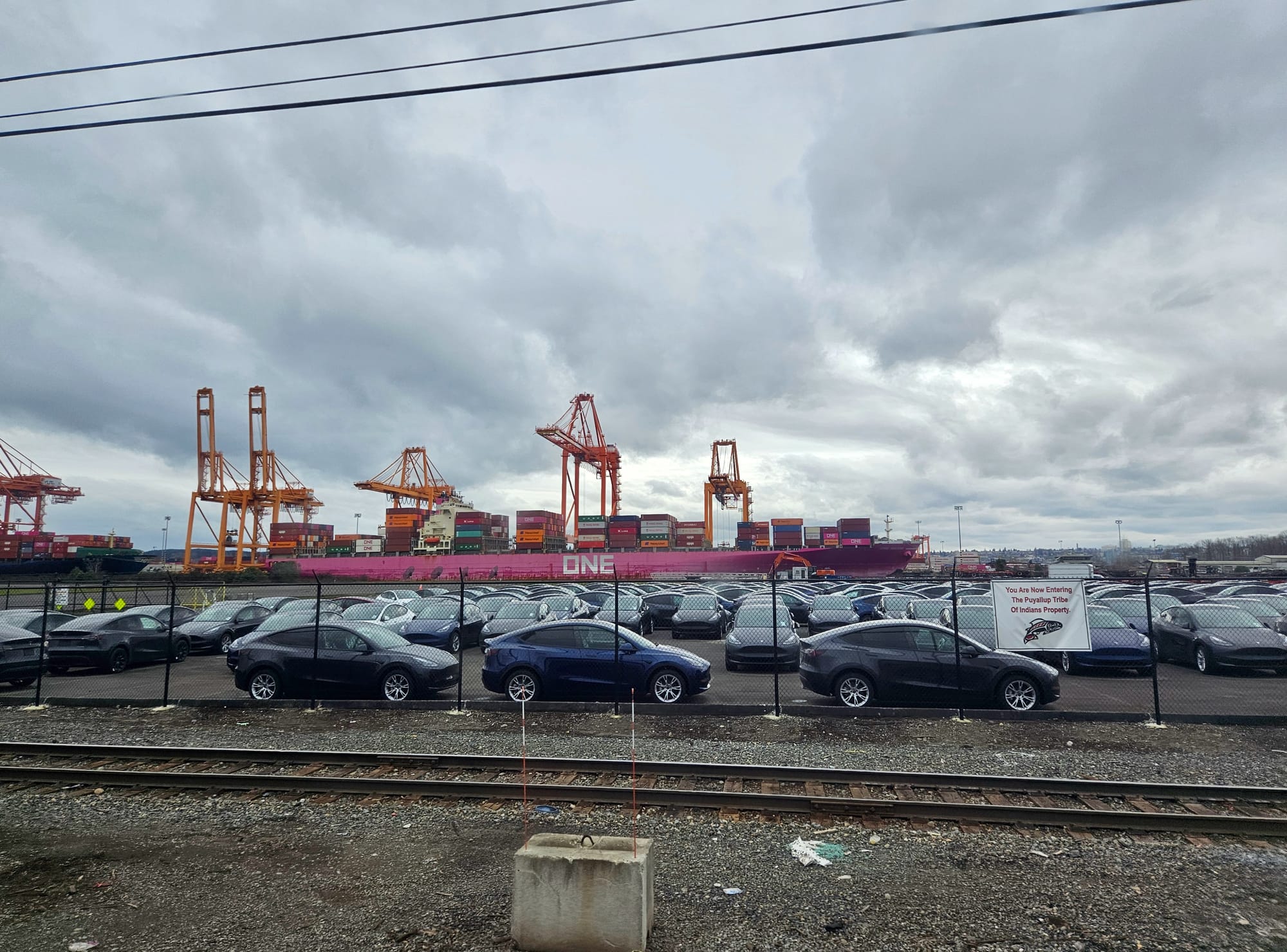 A parking lot full of cars with a magenta container ship partially-loaded with containers in the background.