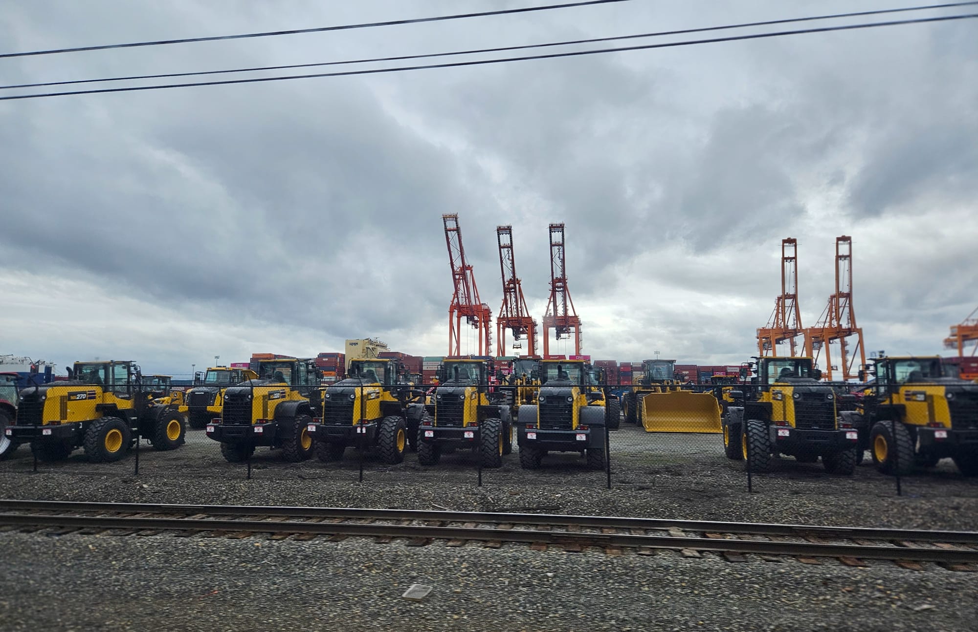 Large yellow tractors fill a parking lot with cranes behind and railroad tracks in the foreground.