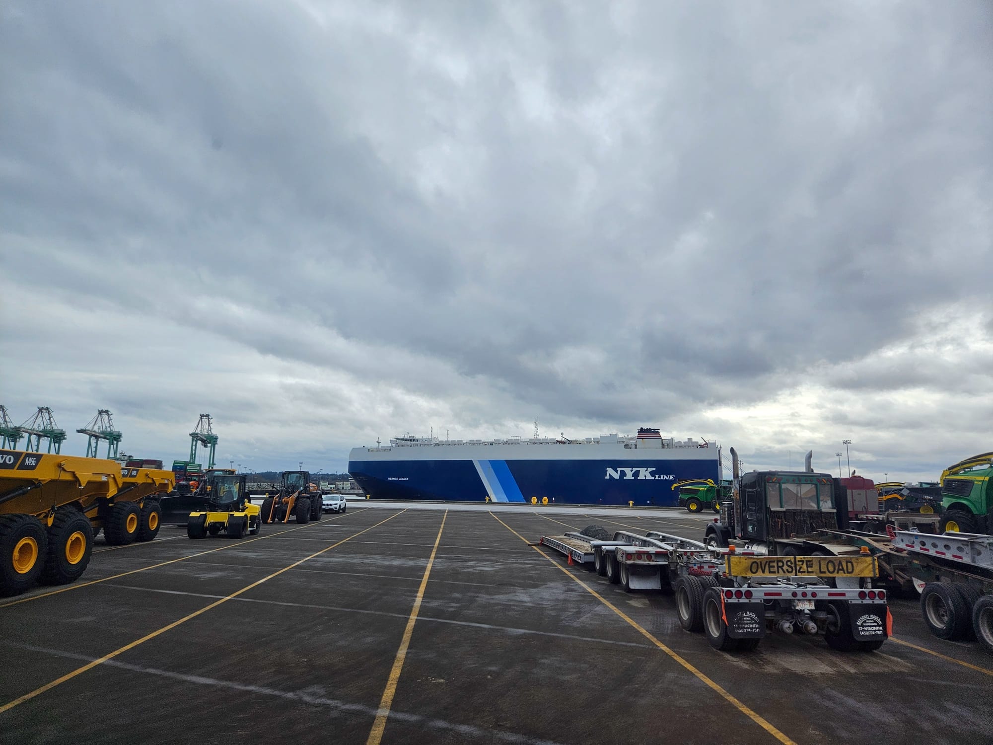 A large and tall blue ship at dock, in the foreground is a parking lot with various vehicles.