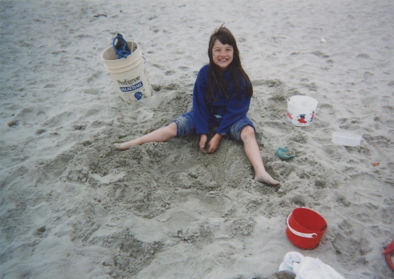 A young girl with long dark hair smiles sitting in the sand with buckets nearby.