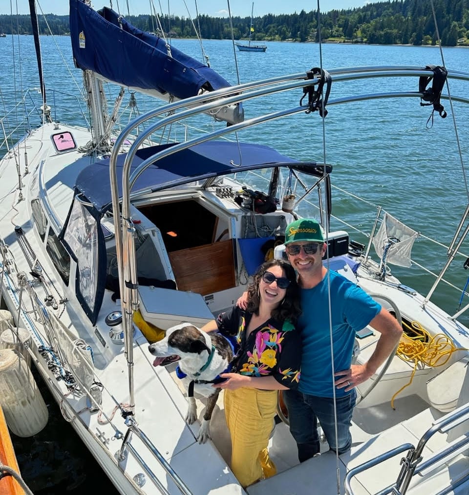 A dog, woman and man pose in the cockpit of a sailboat with blue waters and tree covered shores.