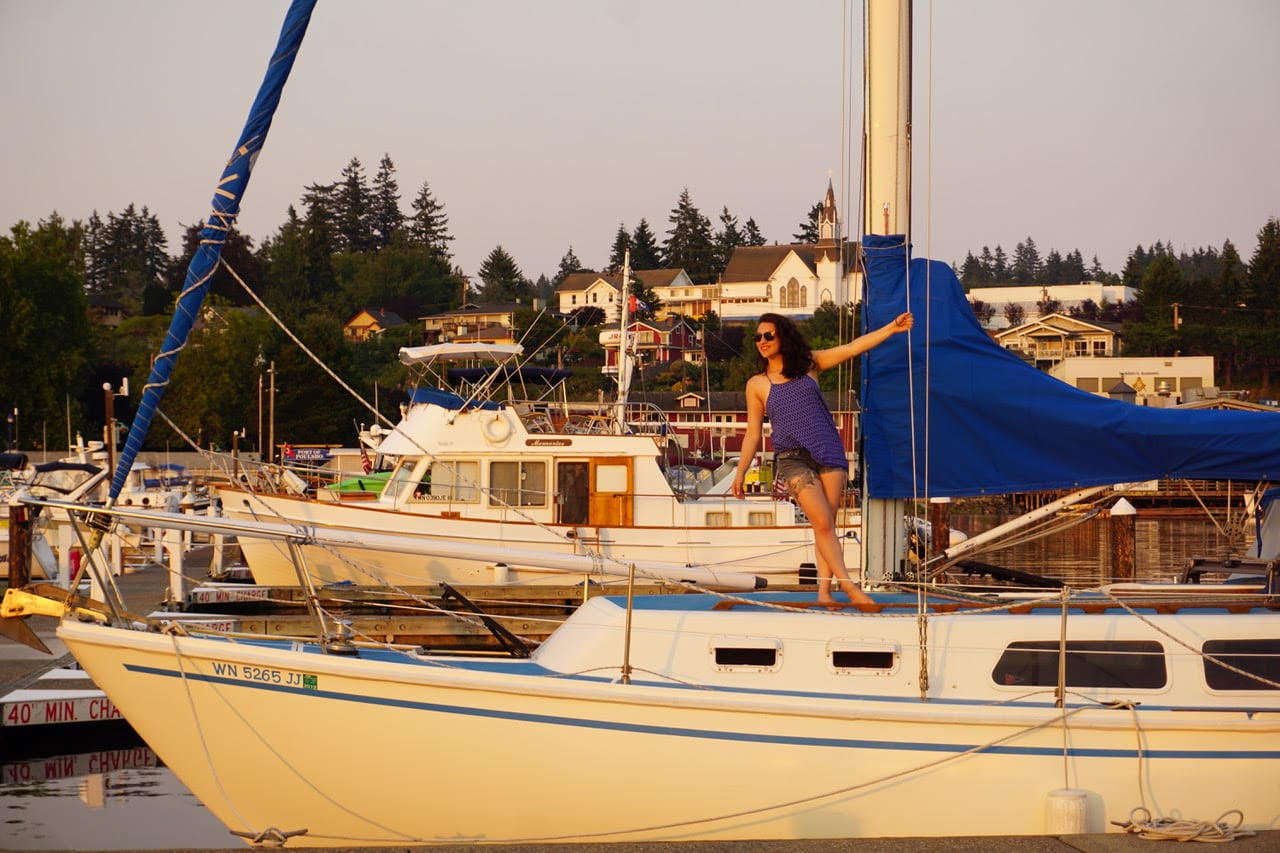 A woman poses on the deck of a sailboat with a blue cover at a dock with buildings behind.