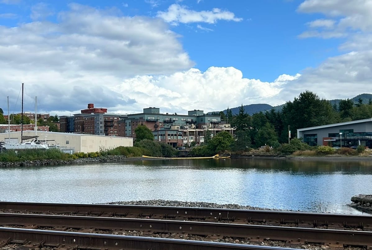 A yellow barrier floats on a small body of water surrounded by buildings and railroad tracks.