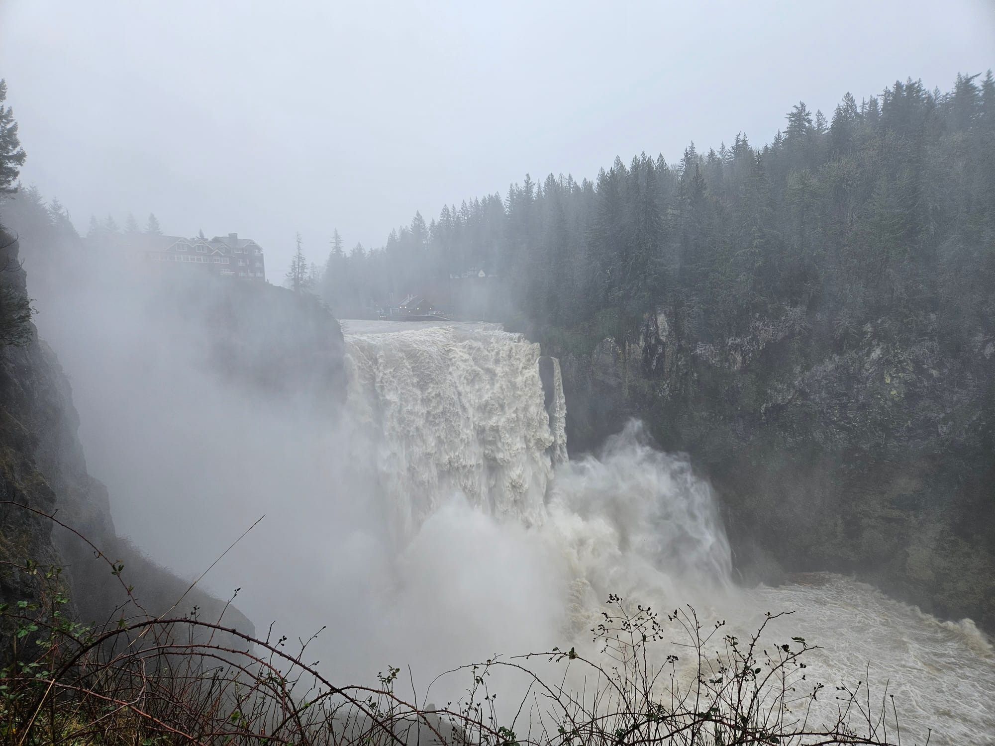 A large slightly brown waterfall with spray surrounded by trees and one building.