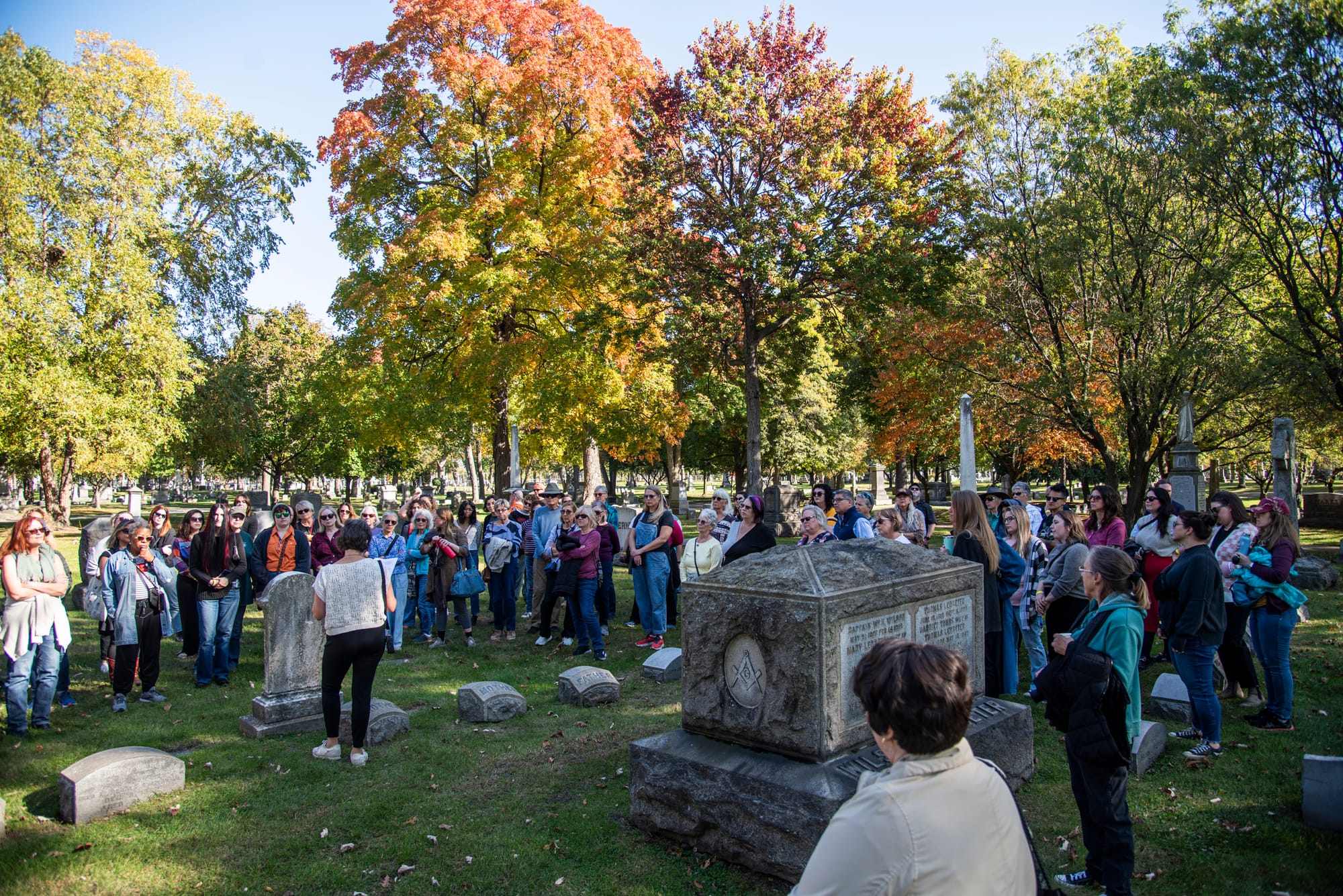 Dozens of people listen to someone speak in a cemetery surrounded by trees changing color.
