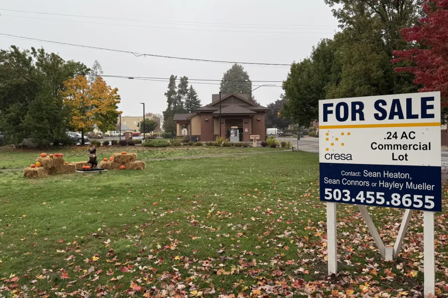A lot with a for sale sign, a statue surrounded by hay bales and a brown building in the back.