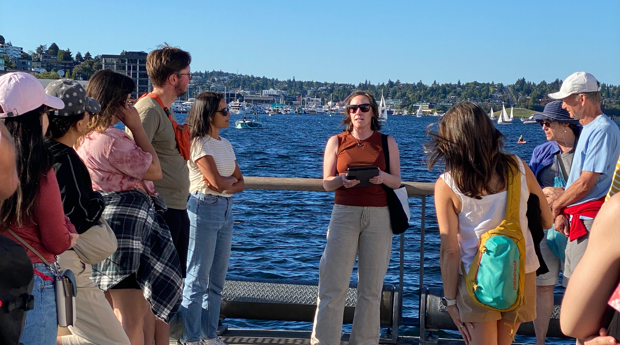 A woman standing in front of a lake speaks to a group of people on a sunny day.