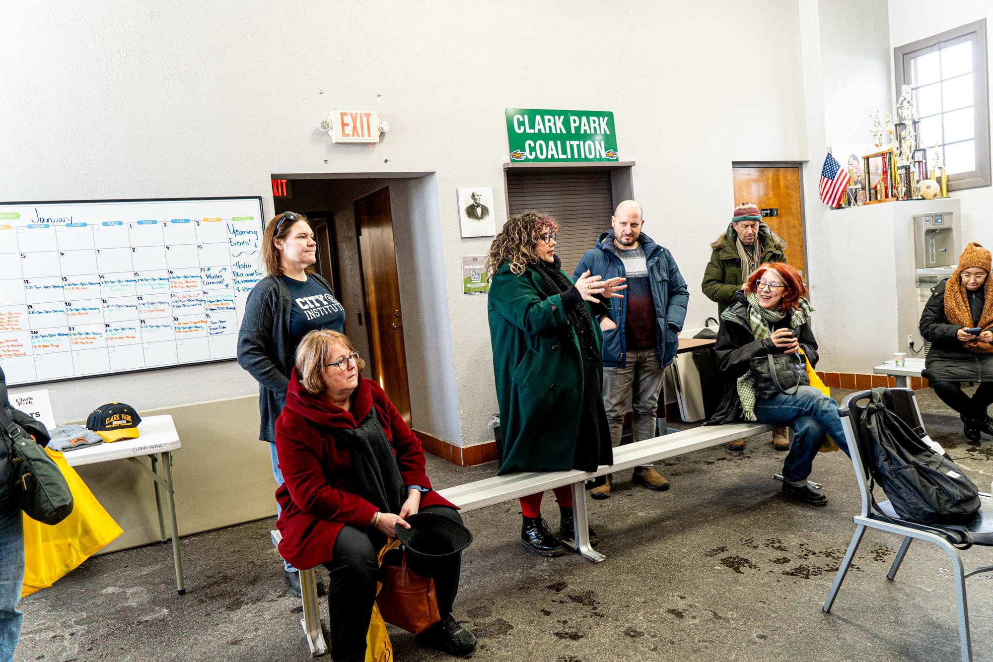 Four people stand and three people sit on benches in a room with a "Clark Park Coalition" sign.