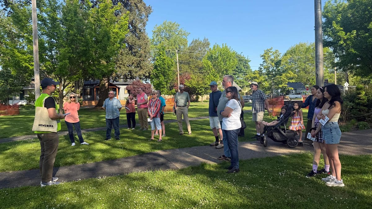 A man in a baseball cap and yellow vest faces more than a dozen people on a park lawn.