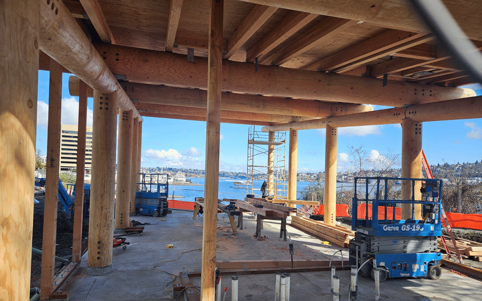 A log framed building under construction with a seaplane on the lake in the distance.