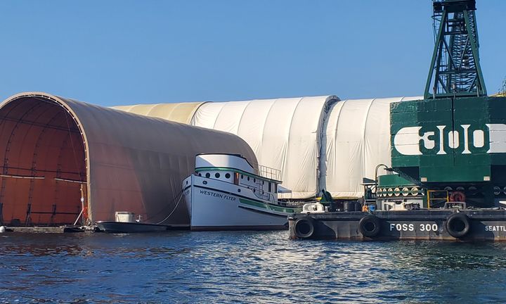 A white boat with green trim with "Western Flyer" painted on the bow docked between large work tents and a crane.