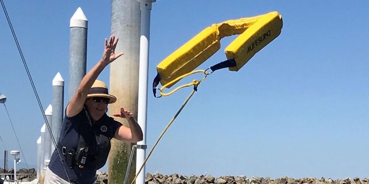 A woman wearing a life jacket and hat throws a yellow, u-shape object with a yellow rope attached from a dock.