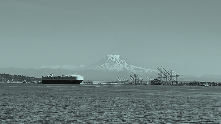 A blue-tinted photo of a waterway with a cargo ship, cranes and a sailboat. A large mountain is in the background.