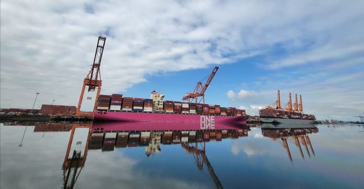 Two ships filled with containers are docked below cranes and reflected in calm water.