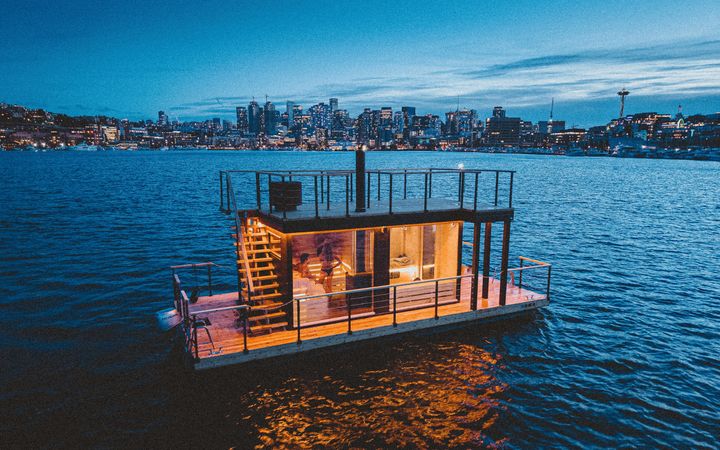 A floating rectangle structure with lights and a deck with the Seattle skyline behind.