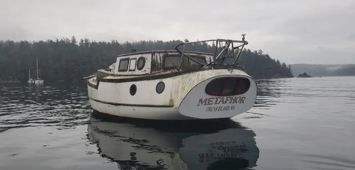 A weathered sailboat with no mast floats in front of a tree-covered island.
